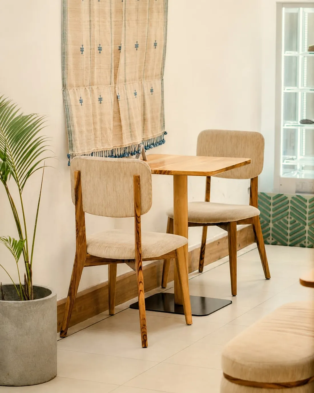 Two cream upholstered chairs at a timber table inside, a woven wall hanging above and green chevron tiles at the skirting.