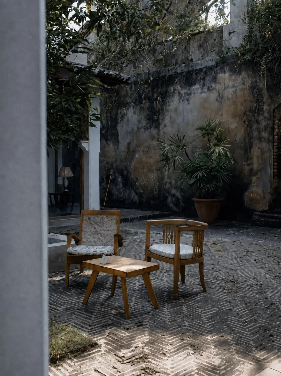 Two timber chairs and a low table in the back courtyard, dappled light across a weathered fort wall.